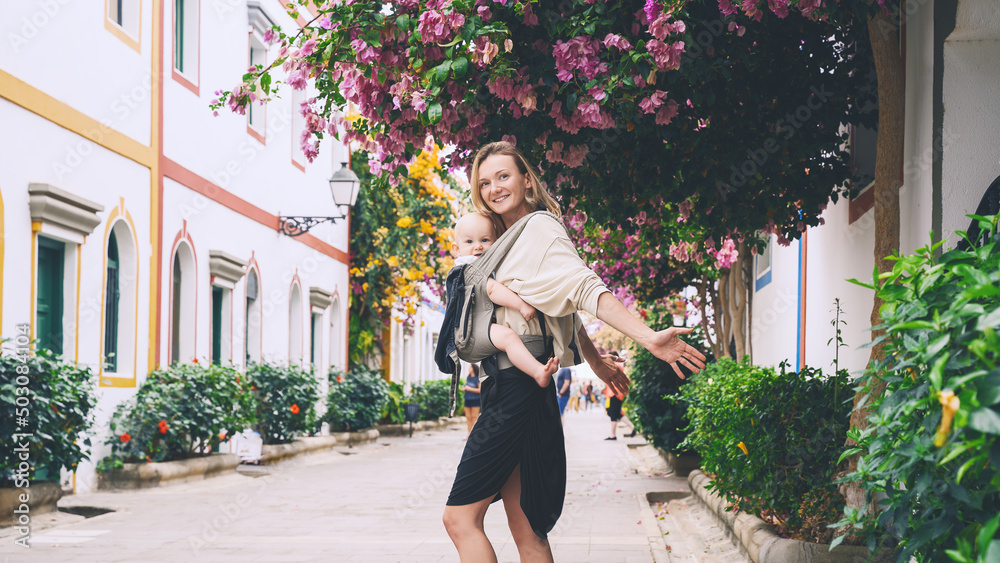 Mother and baby on beautiful street full of blooming trees in Puerto Mogan town, Gran Canaria, Spain. Active travel family vacation in Canary Islands. Woman with child journey in Europe.