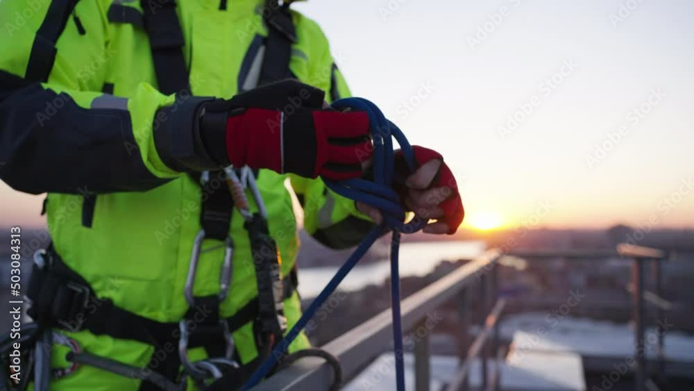 Industrial climber in green outfit ties node with loop attaching carabiner on safety rope and fastens to railing. Man prepares to climb down wall of skyscraper closeup