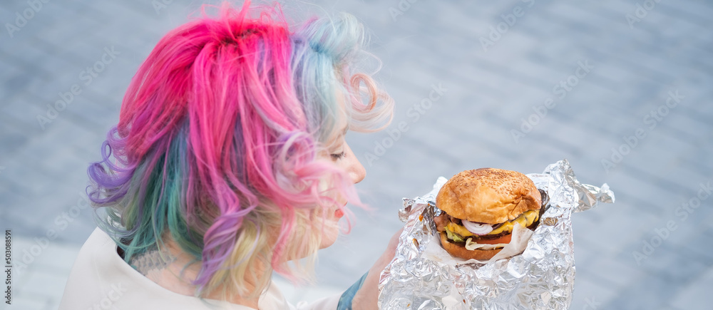 Naklejka premium Caucasian woman with curly colored hair eating burger. Bad eating habits and love of fast food