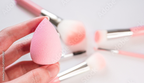 Woman's hand holding a clean makeup sponge with blurred make up brushes in the background