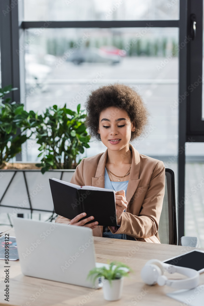 Positive african american businesswoman holding notebook near devices and headphones in office. Positive african american businesswoman holding notebook near devices and headphones in office.