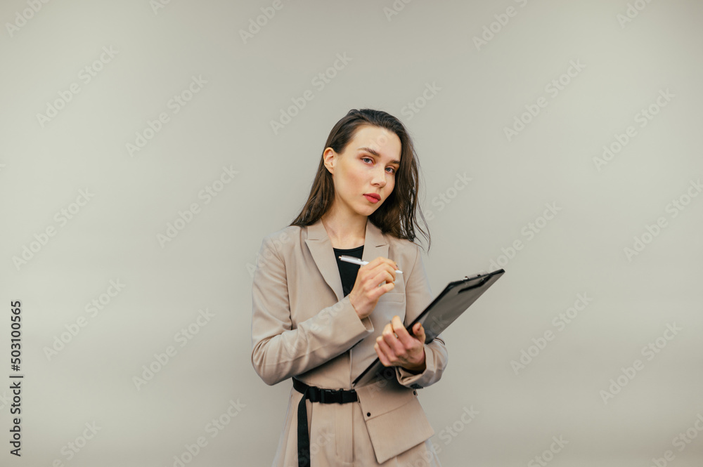 Serious business woman in a jacket stands with a paper tablet and a pen in his hands and looks at the camera on a beige background. Serious business woman in a jacket stands with a paper tablet and a pen in his hands and looks at the camera on a beige background.