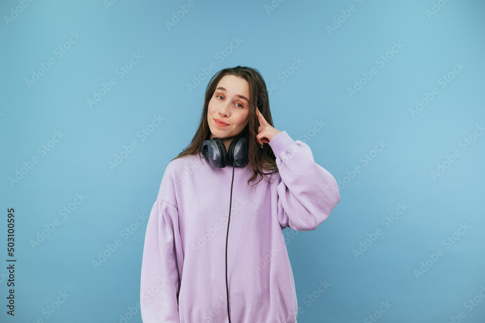 Beautiful woman in headphones and casual clothes points her finger at her temple and looks at the camera with a smile on her face on a blue background. Beautiful woman in headphones and casual clothes points her finger at her temple and looks at the camera with a smile on her face on a blue background.