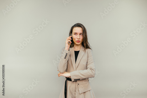 Beautiful woman in a jacket stands on a beige background and talks on the phone with a serious face and looks away.