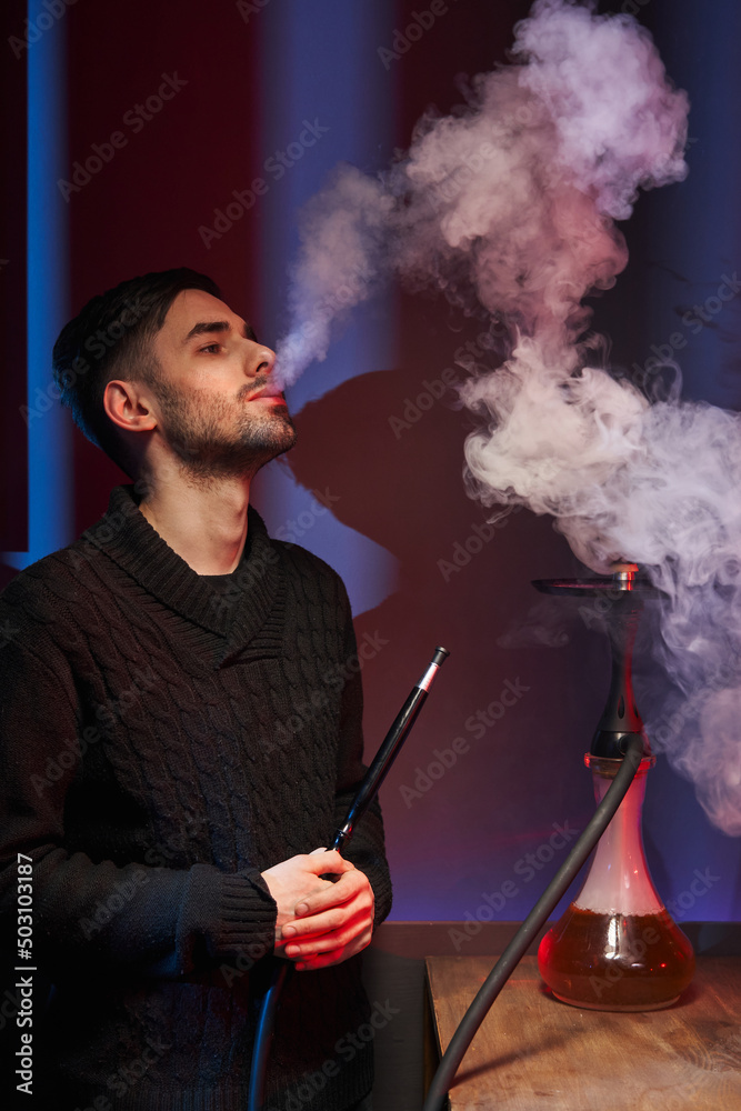 a young man smokes a hookah, releasing a large cloud of smoke from his mouth. next to it is a hookah with a transparent flask. atmospheric cafe with hookah