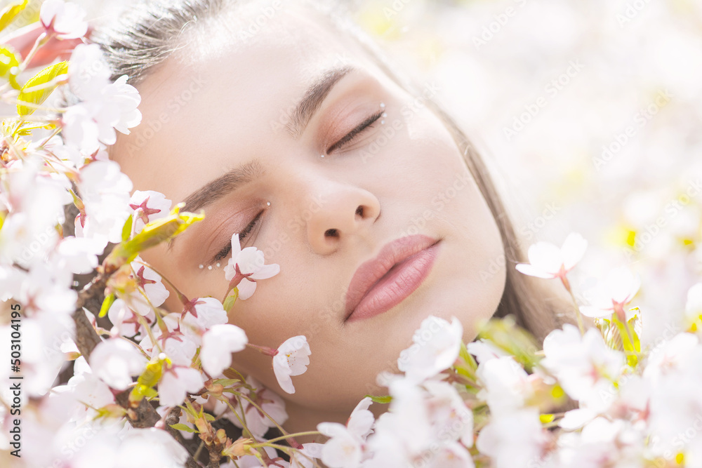 Beauty, lifestyle and nature concept. Beautiful brunette woman portrait in white sakura blossom background during spring time in sunny day. Model placed head on blossoms with closed eyes and dreaming
