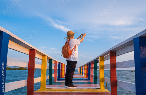 Rear side view of Asian female tourist photographing with smartphone on rainbow wooden bridge at sea viewpoint against clouds on blue sky background in evening time