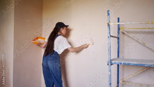 Professional painter female paints the wall with roller and tray on construction site. Young woman in overalls and cap doing home decoration and renovetion. Worker makes DIY repair work in the room. photo