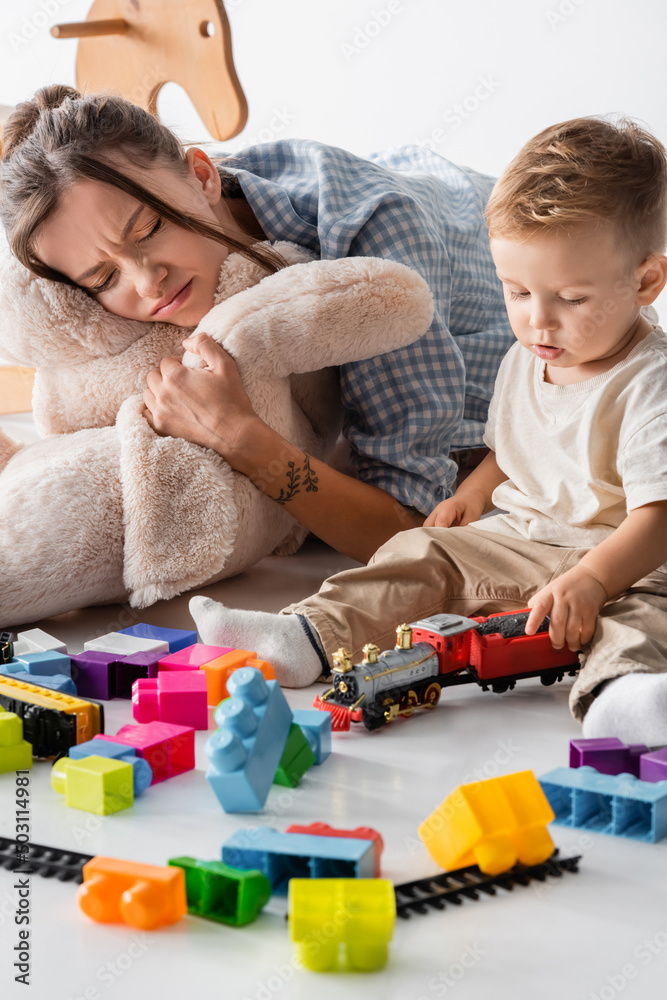 frowning woman hugging soft toy near toddler son playing on white. frowning woman hugging soft toy near toddler son playing on white.