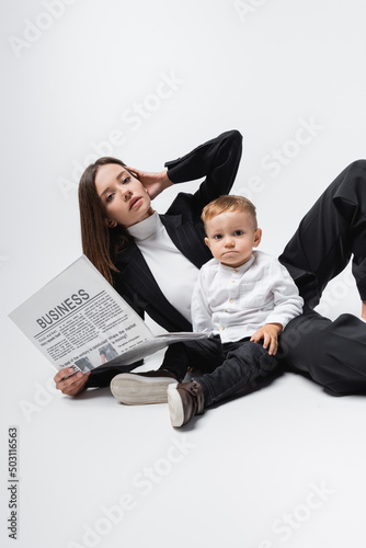businesswoman with newspaper looking at camera near little son on white.