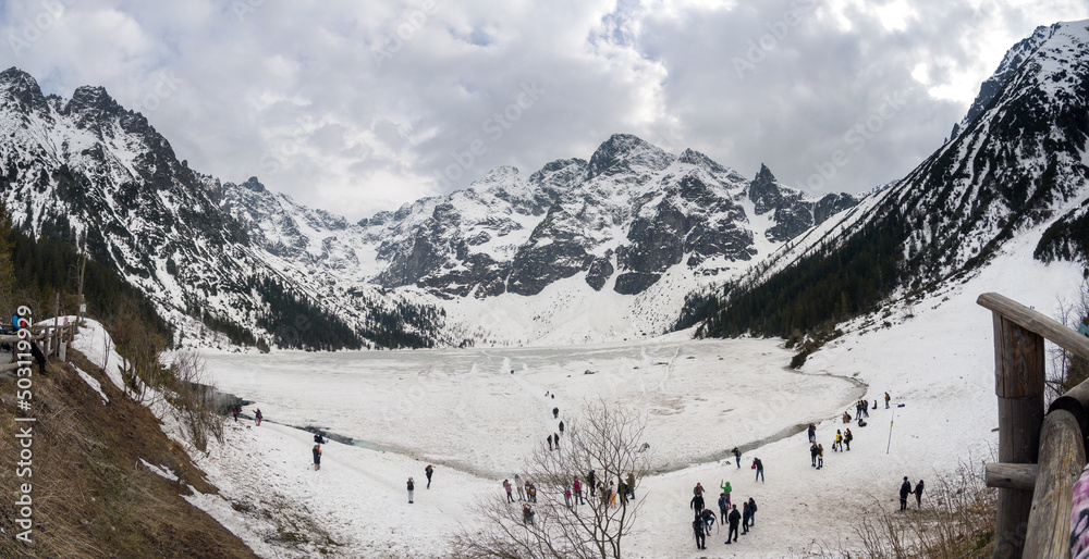 Zakopane, Poland - May 04, 2022: Panorama of Morskie oko frozen lake covered with snowy tatra mountains with people or tourists walking on snow - obrazy, fototapety, plakaty 