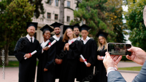 Graduation day for a group of multiracial graduates students posing with a large smile very excited in front of the smartphone in the college park one of the parents old man taking pictures for