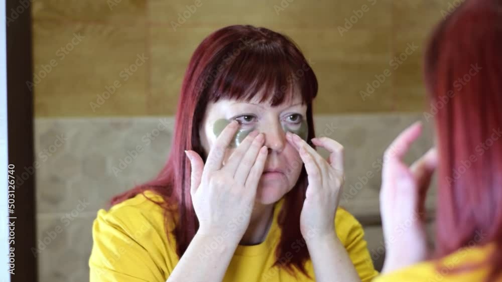 Middle-aged red-haired woman is applying under eye patches standing in front of the mirror in the bathroom. Beauty skin care routine. Morning self-care Middle-aged red-haired woman is applying under eye patches standing in front of the mirror in the bathroom. Beauty skin care routine. Morning self-care