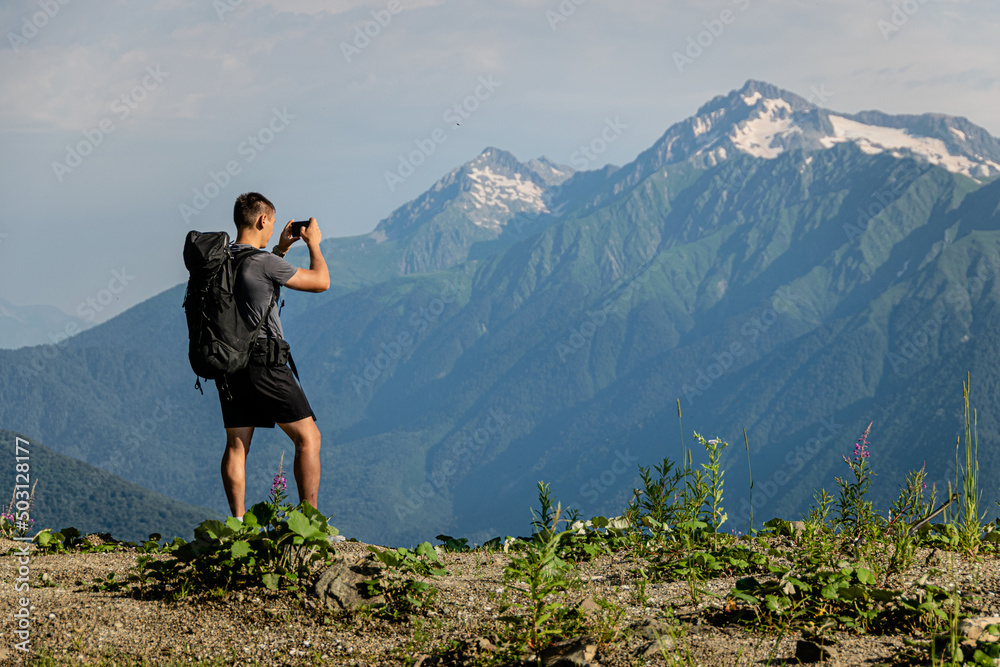 A male hiker with a large backpack photographs a mountain landscape