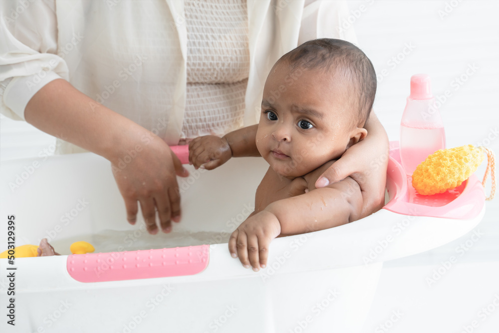 Happy adorable African newborn baby bathing in bathtub. Mother washing her little daughter in warm water with yellow sponge and shower gel. Newborn baby cleanliness care concept Happy adorable African newborn baby bathing in bathtub. Mother washing her little daughter in warm water with yellow sponge and shower gel. Newborn baby cleanliness care concept