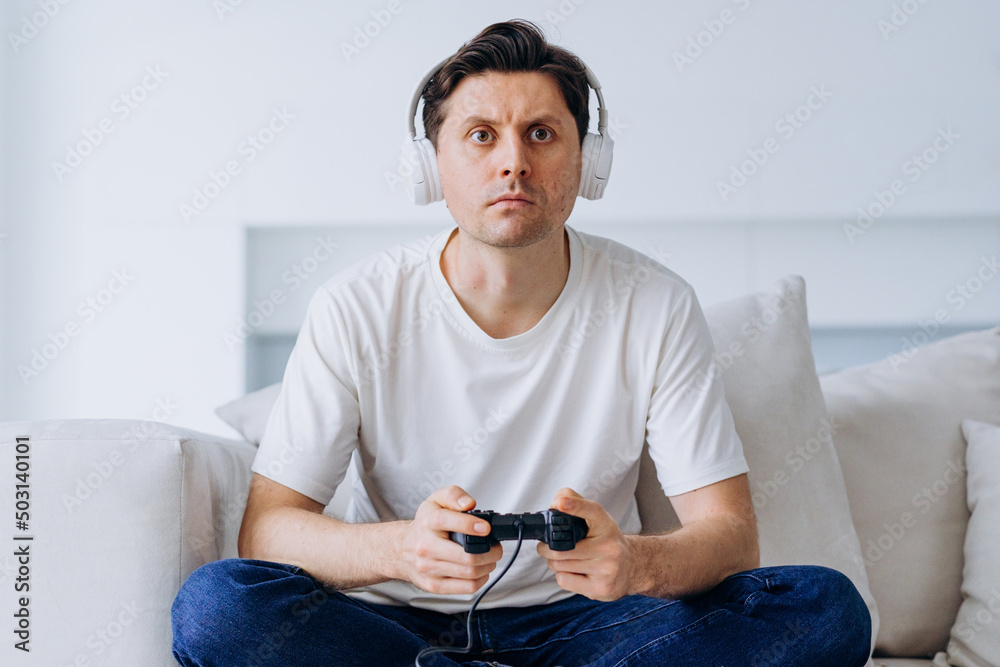 Excited man in wireless headphones sits on sofa in living room plays non-stop catch-up game with controller and loses illuminated by screen light Excited man in wireless headphones sits on sofa in living room plays non-stop catch-up game with controller and loses illuminated by screen light