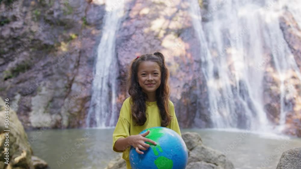 Smily girl stands with a globe and points to it in front of a waterfall. The concept of World Environment Day, pollution, Global Warming, Earth day. Smily girl stands with a globe and points to it in front of a waterfall. The concept of World Environment Day, pollution, Global Warming, Earth day.
