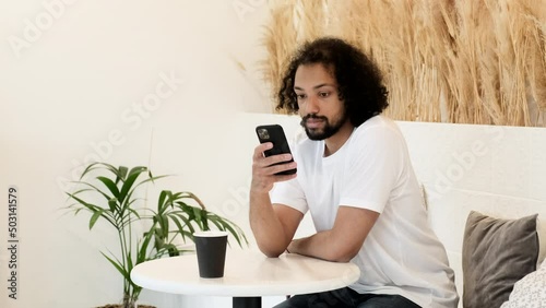 A Young Indian Man Looks Thoughtfully, Works on A New Online Project, Uses A Smartphone, Is in A Cafe