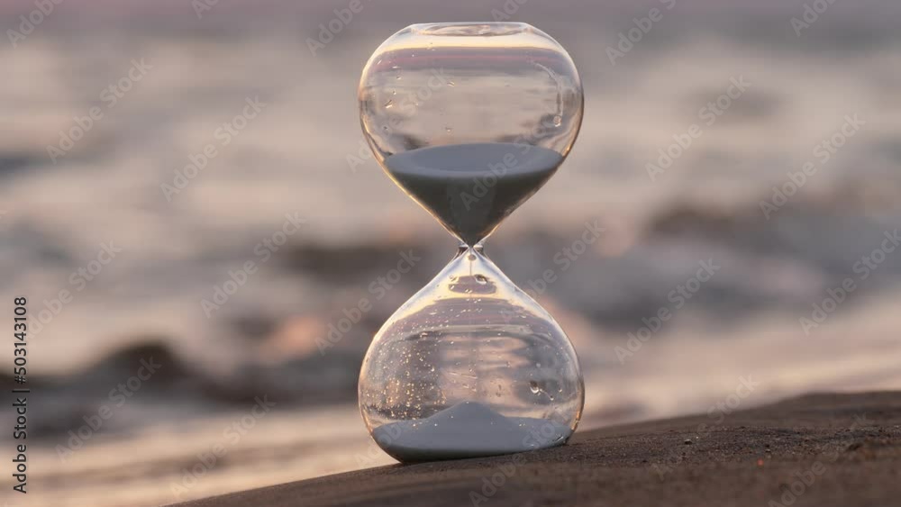 Hourglass stand on sea beach at evening, sometimes waves wash bottom and water drops reach glass. Selective focus, blurred waves on background. Dim natural light