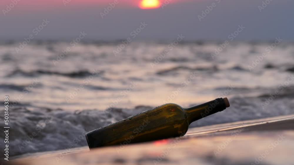 Bottle with message thrown ashore lies and then swayed from splash of oncoming wave. Close up shot of glass bottle traveled across sea to unknown beach, blurred sunset visible on background