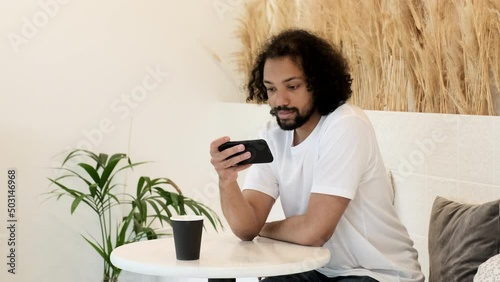 Asian man looking at smartphone screen watching online lesson and movie while sitting on couch and doing meeting online. Indian guy watching online videos, working from home concept