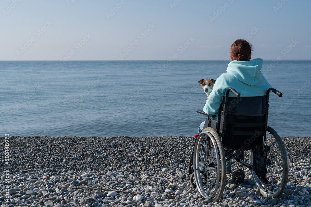 Caucasian woman in a wheelchair cuddling with a dog near the sea. 