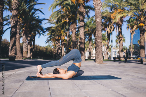 Young fit girl in tracksuit clothes stretching body muscles during daytime workout near palm trees, Asian female yogi spending daytime for holistic healing on hatha retreat practice in city © BullRun