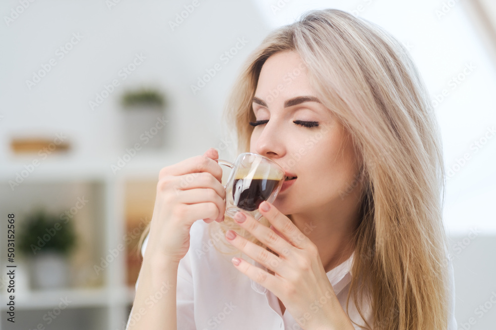 A young beautiful Caucasian blonde woman rests at home. A woman sits at a table, drinking coffee from a glass cup and reading a magazine. A young beautiful Caucasian blonde woman rests at home. A woman sits at a table, drinking coffee from a glass cup and reading a magazine.