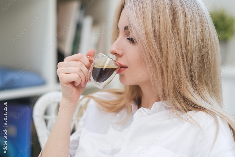 A young beautiful Caucasian blonde woman rests at home. A woman sits at a table, drinking coffee from a glass cup and reading a magazine. A young beautiful Caucasian blonde woman rests at home. A woman sits at a table, drinking coffee from a glass cup and reading a magazine.