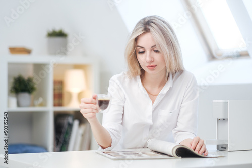 A young beautiful Caucasian blonde woman rests at home. A woman sits at a table  drinking coffee from a glass cup and reading a magazine.
