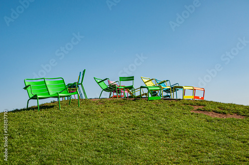 Valokuvatapetti Colorful chairs on the green hillside against the gradient blue sky