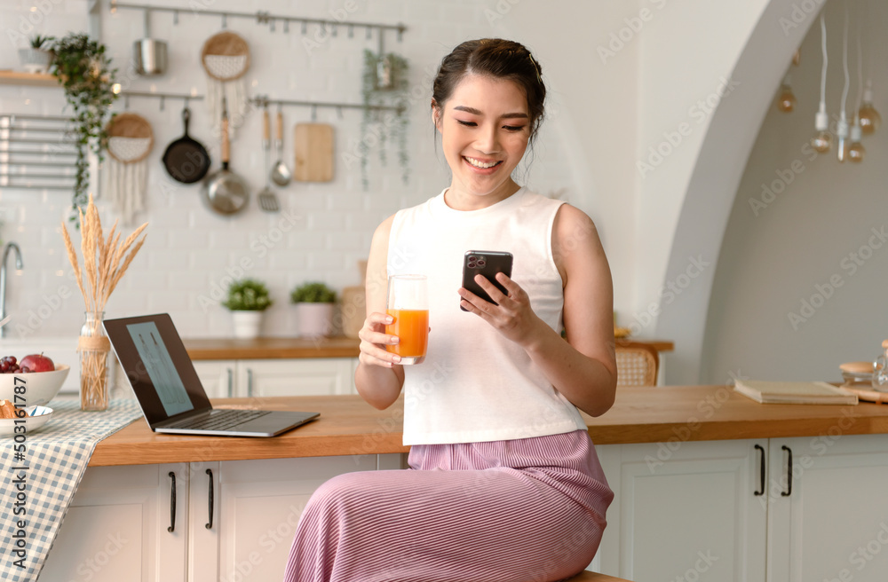 Young woman Asian happy rest drinking orange juice and using mobile phone in kitchen room. Young woman Asian happy rest drinking orange juice and using mobile phone in kitchen room.