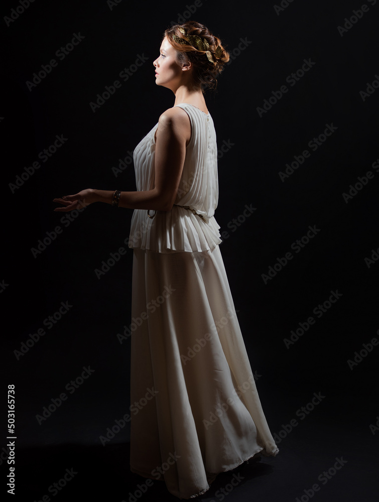Fototapeta premium An ancient heroine, a young woman in the image of an ancient Greek goddess or muse. A noble heroine in a white tunic and a laurel wreath, a full-length photo on a black background
