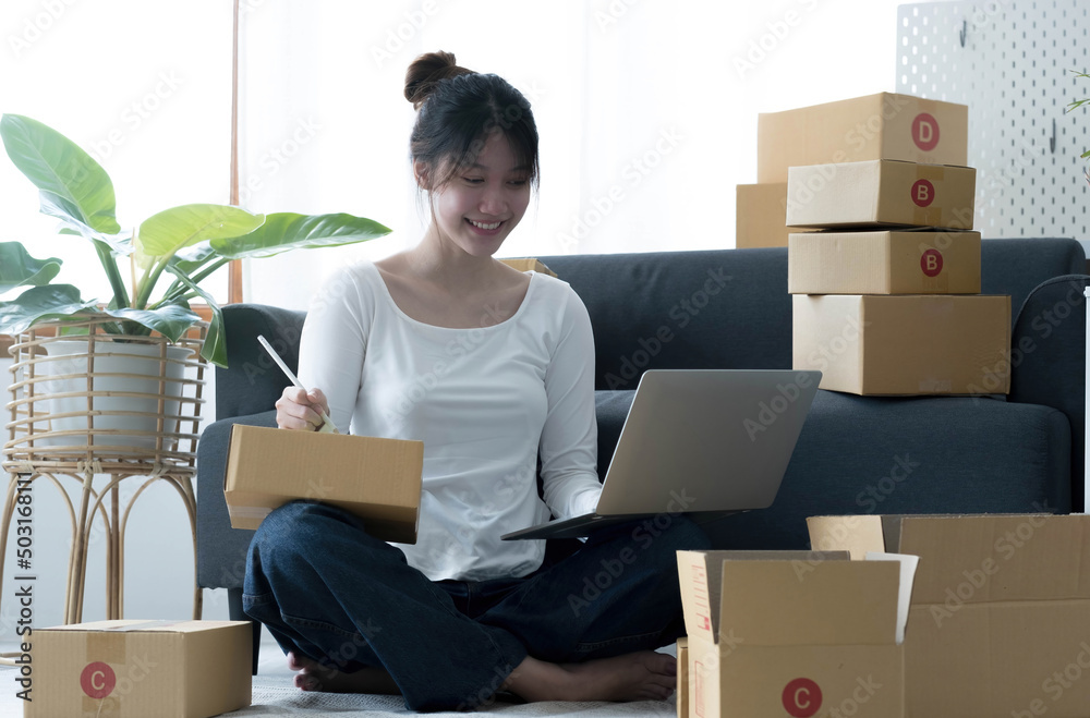 Fototapeta premium Smiling young Asian business owner woman prepare parcel box and standing check online orders of product for deliver to customer on laptop computer. Shopping Online concept.