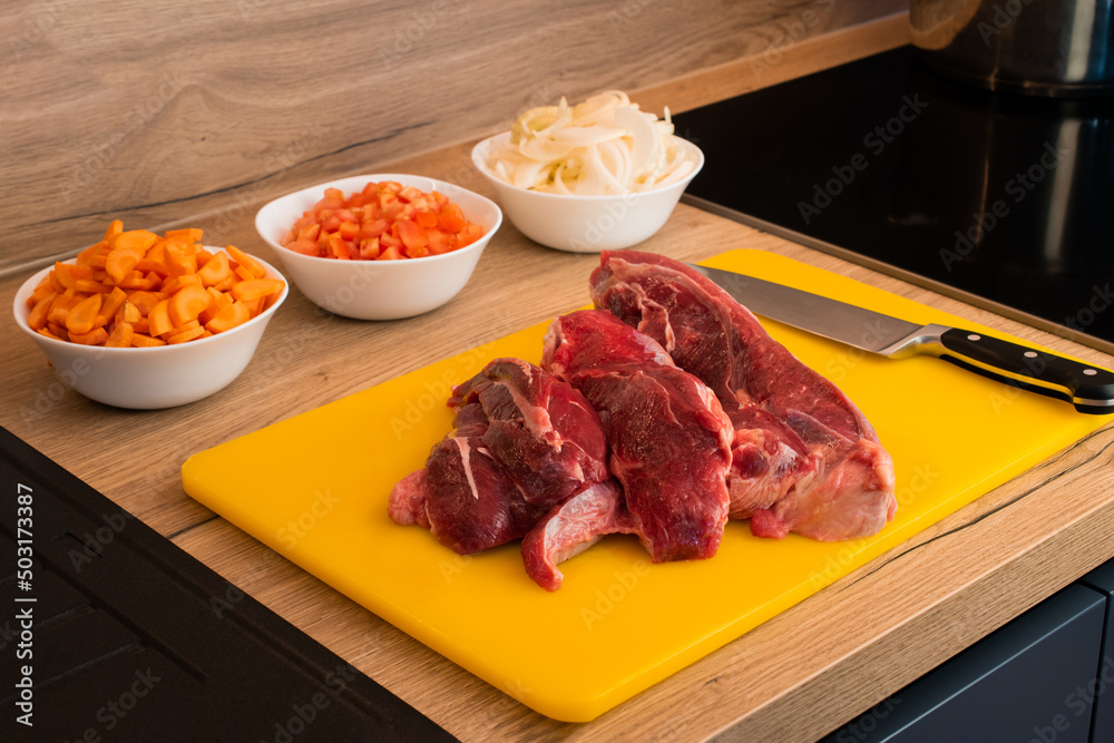 Large pieces of fresh beef laid out on a cutting board, plates with cut onion, paper and carrot. The process of cooking Large pieces of fresh beef laid out on a cutting board, plates with cut onion, paper and carrot. The process of cooking