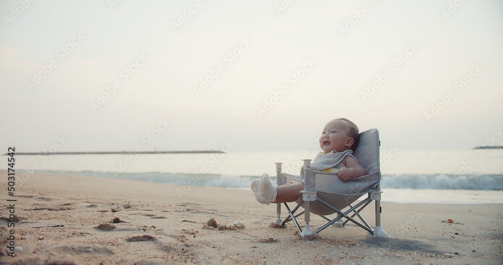 happy cute adorable Asian baby infant sitting relaxing on little chair and smiling with waves on background at seaside tropical sandy beach in sunrise during holiday vacation summertime Thailand	
