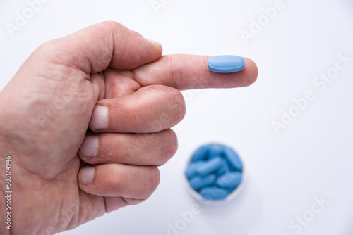 Male hand showing a blue pill on a finger and in the background container full of blue pills