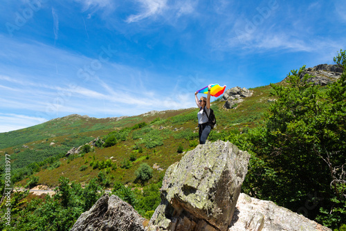A woman with a backpack, perched on a rock on top of a mountain, arms raised holding a rainbow flag waving in the wind.