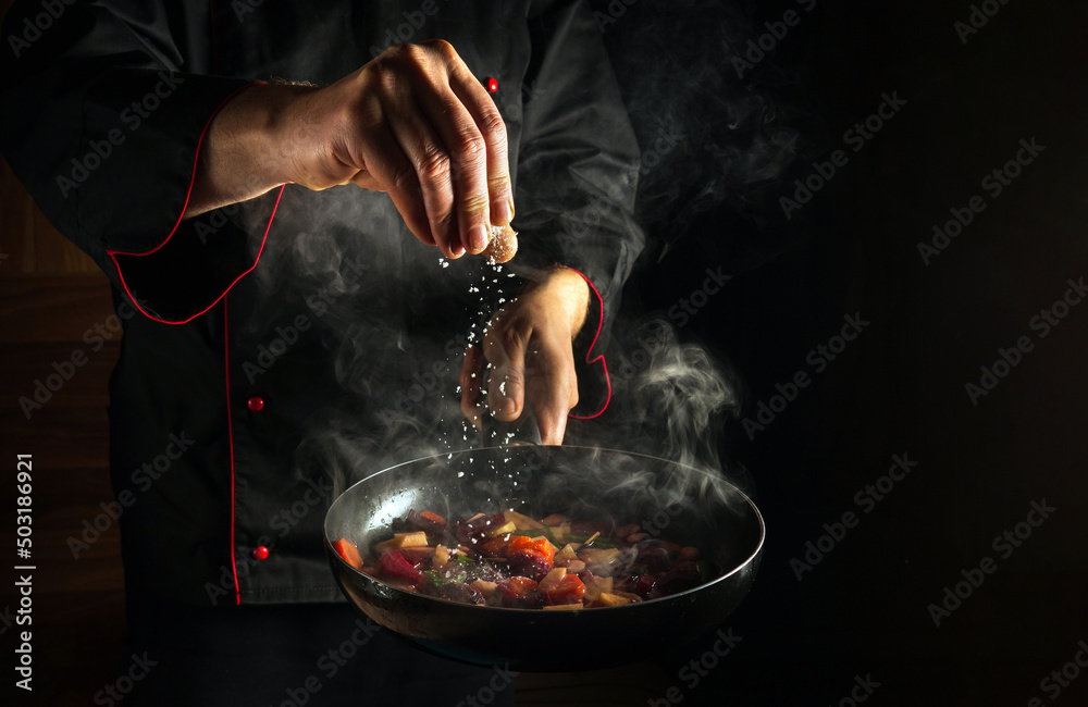 Professional chef adds salt to a steaming hot pan. Grande cuisine idea for a hotel with advertising space. - obrazy, fototapety, plakaty 