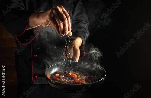 Fototapeta Naklejka Na Ścianę i Meble -  Professional chef adds salt to a steaming hot pan. Grande cuisine idea for a hotel with advertising space.