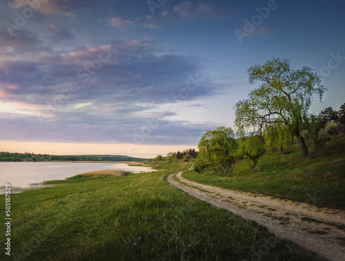 Rural landscape with a country road between lake and forest. Idyllic summer scene, peaceful evening with colorful sunset sky