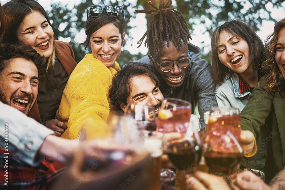 Multiethnic group of excited young people having fun toasting wine and beers together at terrace birthday party - lifestyle concept of multigenerational diverse family stay together - focus center man