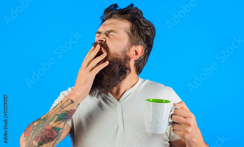 Sleepy bearded man drinking coffee. Yawning guy with mug or cup of hot drink. Morning refreshment. photo