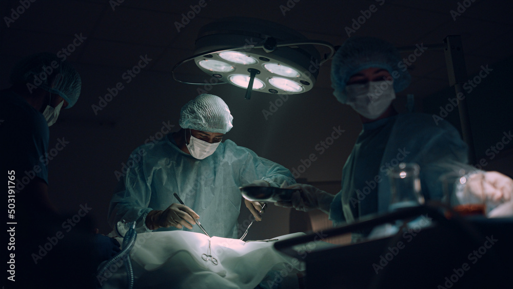 Doctors team performing surgical operation in dark hospital operating room. - obrazy, fototapety, plakaty 