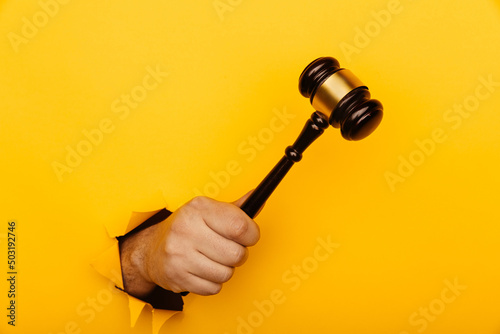 Male hand holding a wooden gavel through torn yellow background. Law and auction aconcept