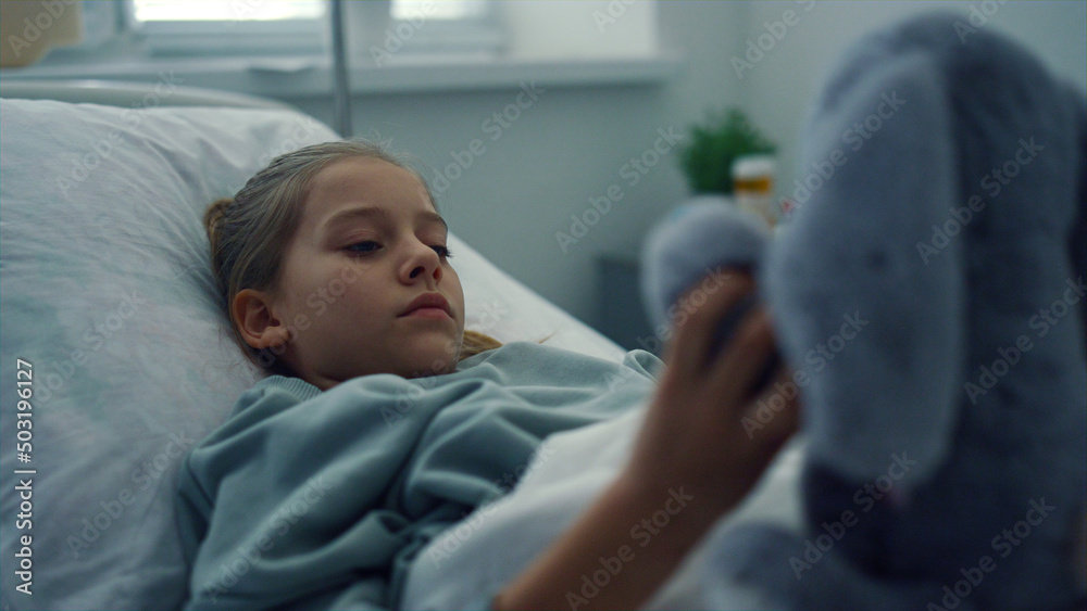 Upset girl lying hospital bed portrait. Sad kid playing with plush toy alone. Upset girl lying hospital bed portrait. Sad kid playing with plush toy alone.