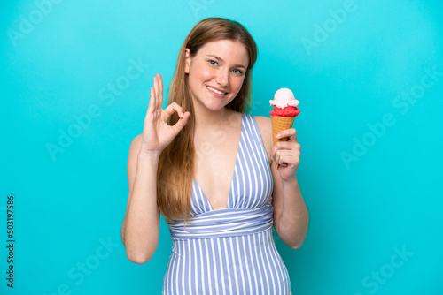 Young caucasian woman in swimsuit eating ice cream isolated on blue background showing ok sign with fingers