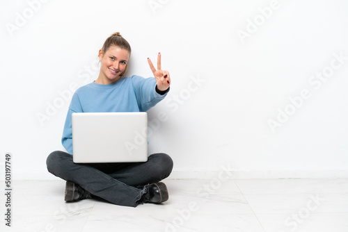 Young woman with a laptop sitting on the floor smiling and showing victory sign © luismolinero