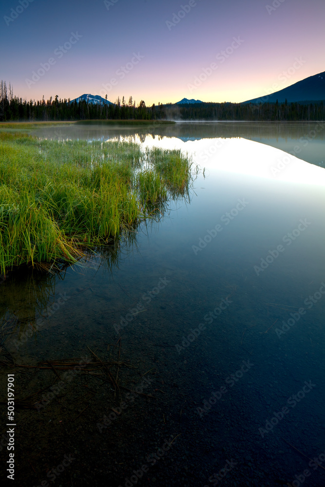 Beautiful mountain lake with bright green grasses and clear clean water reflecting the light of the clear blue sky and a light mist rising of it just before sunrise with Mt Hood in the distance