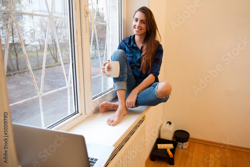 Woman in casual denim clothes sitting on the window sill and drinking coffee